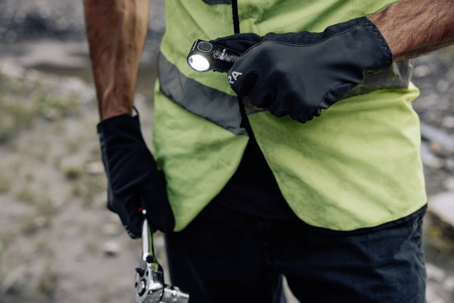 A worker in a high-visibility vest is holding a torch and a tool. The background is blurred, showing a construction site.