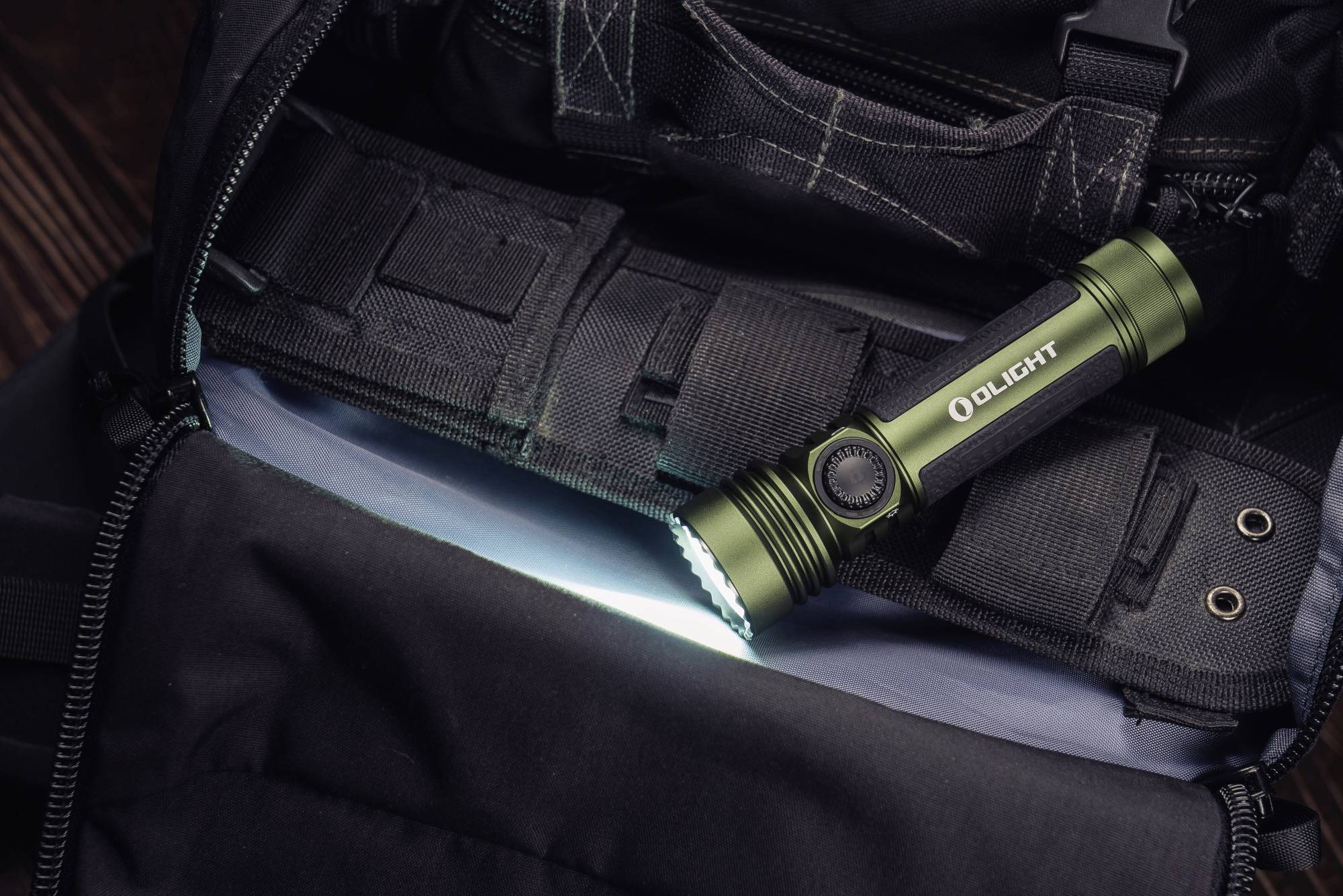 A green torch illuminates an open bag. The torch is resting on multiple compartments and is pointing towards a white surface.