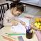 A child is drawing at a table covered with pencils and a notebook; beside it stands a fruit basket with citrus fruits.