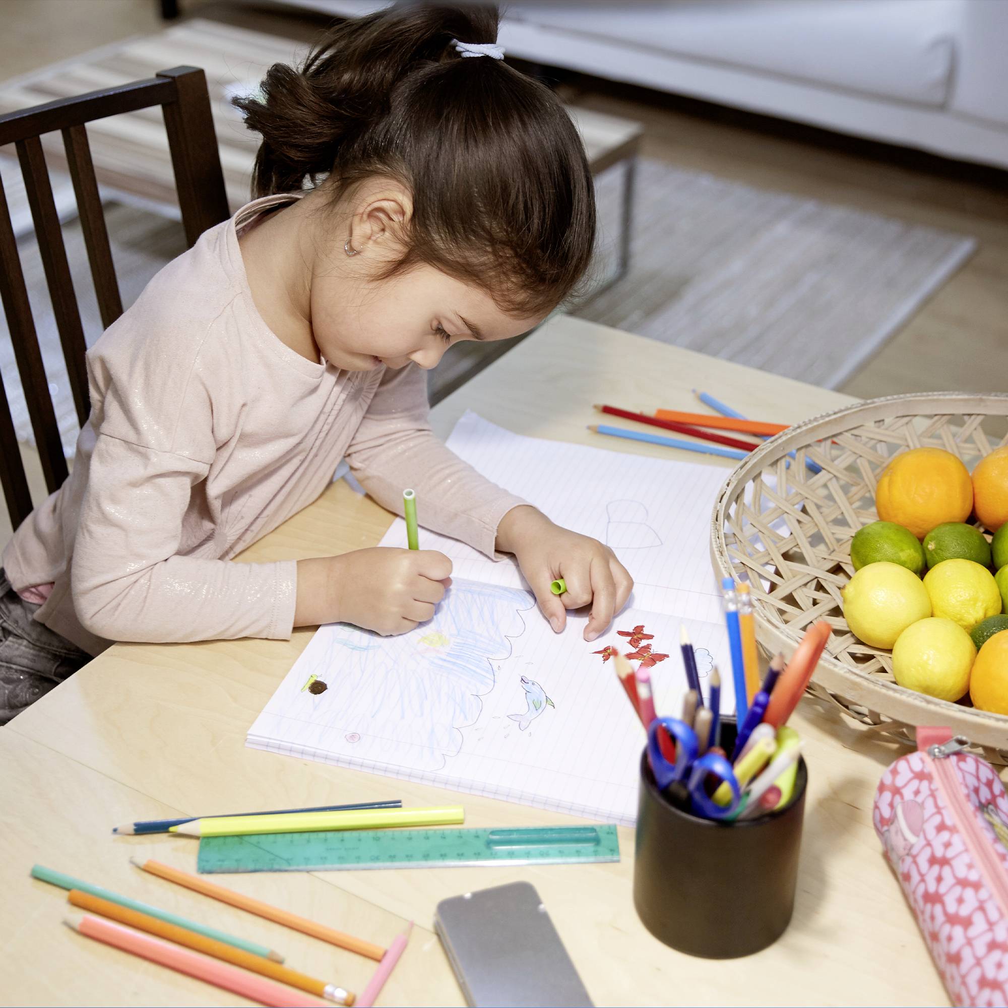 A child is drawing at a table, surrounded by coloured pencils, rulers, and a basket of fruit.