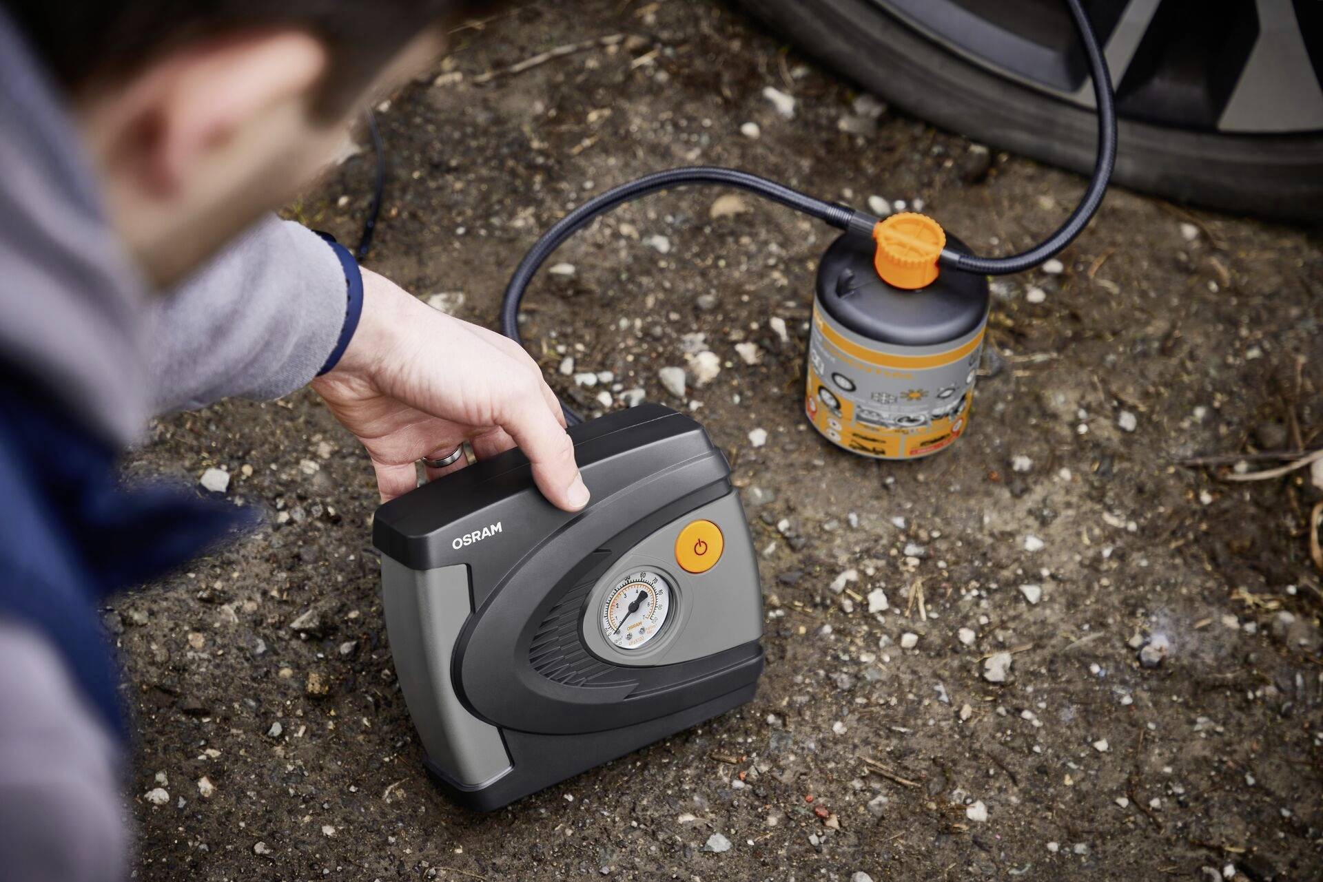 A person is using a portable air compressor to inflate a tyre. The compressor is connected to an orange canister.