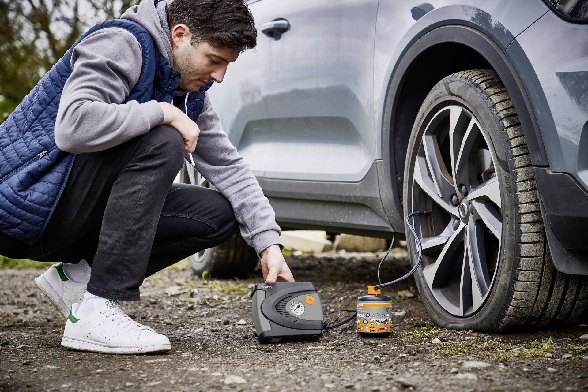 A man is inflating a tyre on a grey saloon car using a portable compressor. The tyre is partially inflated.