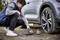A man is inflating a tyre on a grey saloon car using a portable compressor. The tyre is partially inflated.
