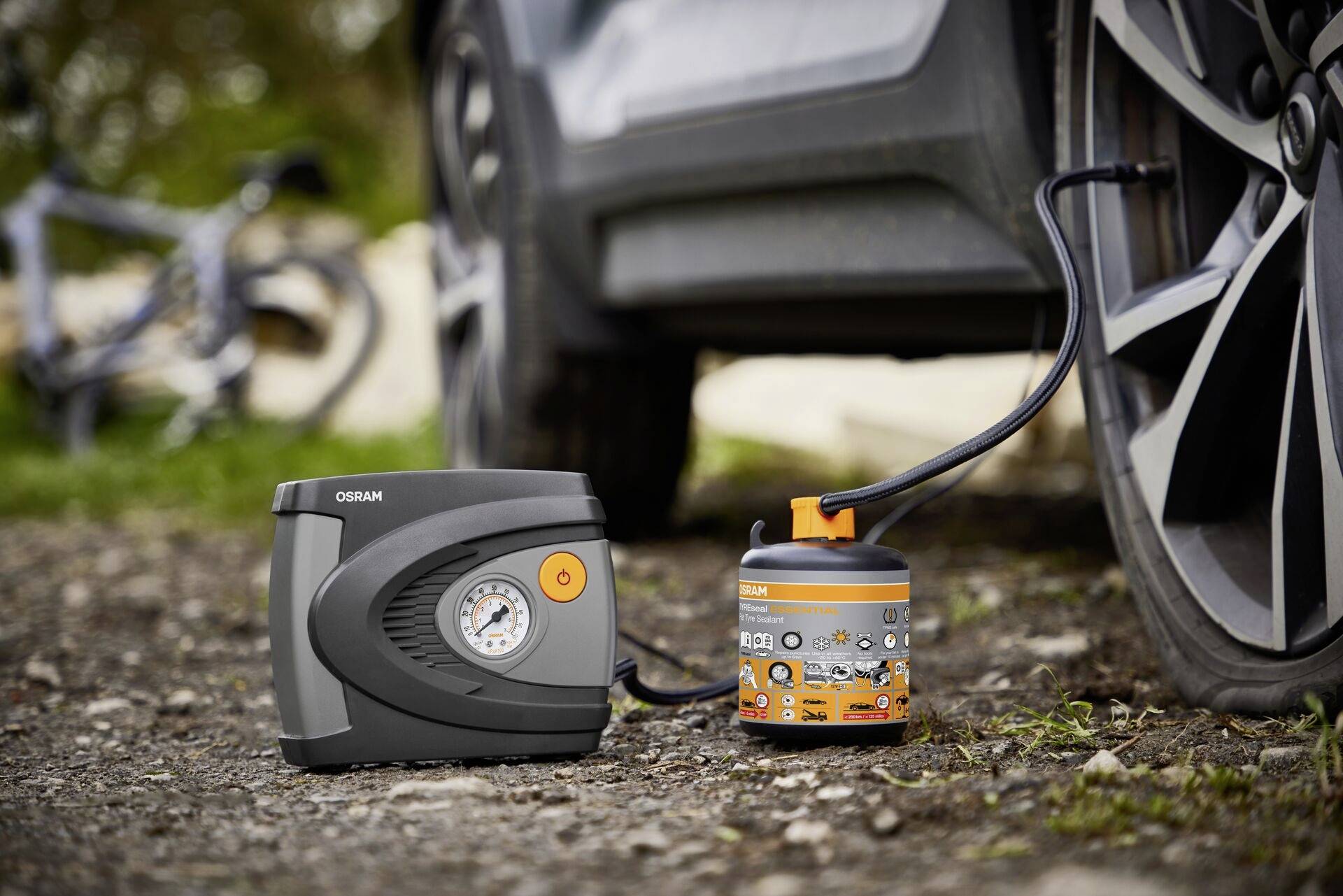 A tyre repair kit next to a car tyre on a gravel road, consisting of a pressure container and an air compressor.