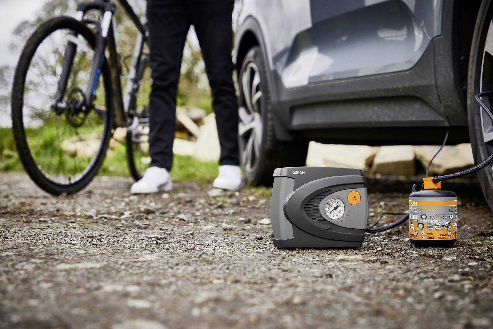 A person is standing next to a car while a tyre is being inflated with a portable compressor. A bicycle is leaning nearby.