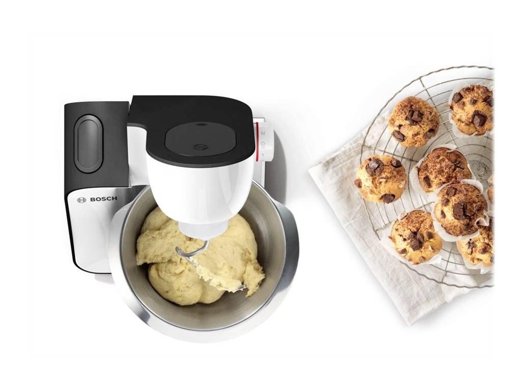 Kitchen mixer with a dough hook kneads dough in a bowl. Freshly baked rolls are placed on a wire rack beside it.