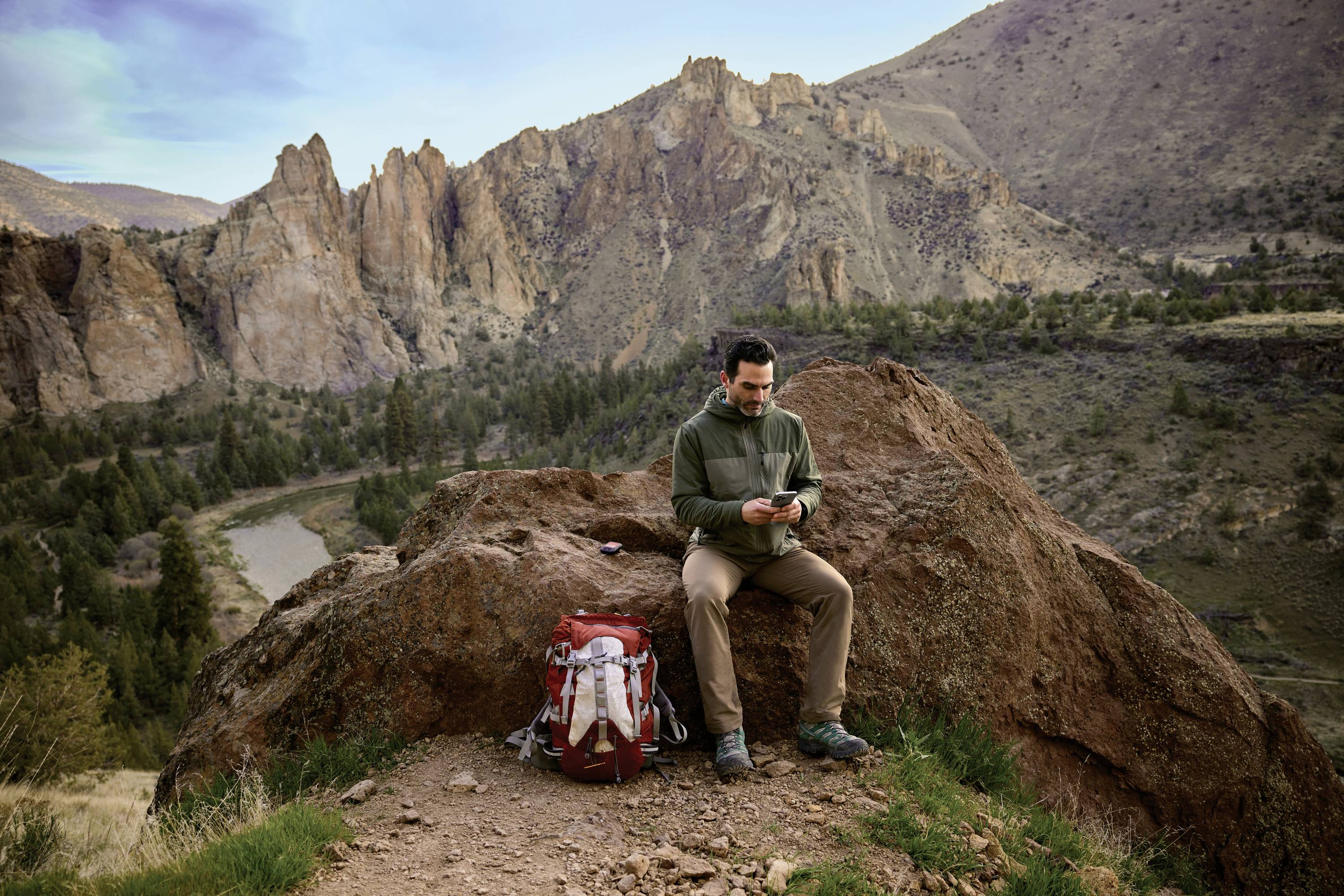 A person sits on a rock in a mountainous landscape, with a red rucksack beside them. A river is visible in the background.