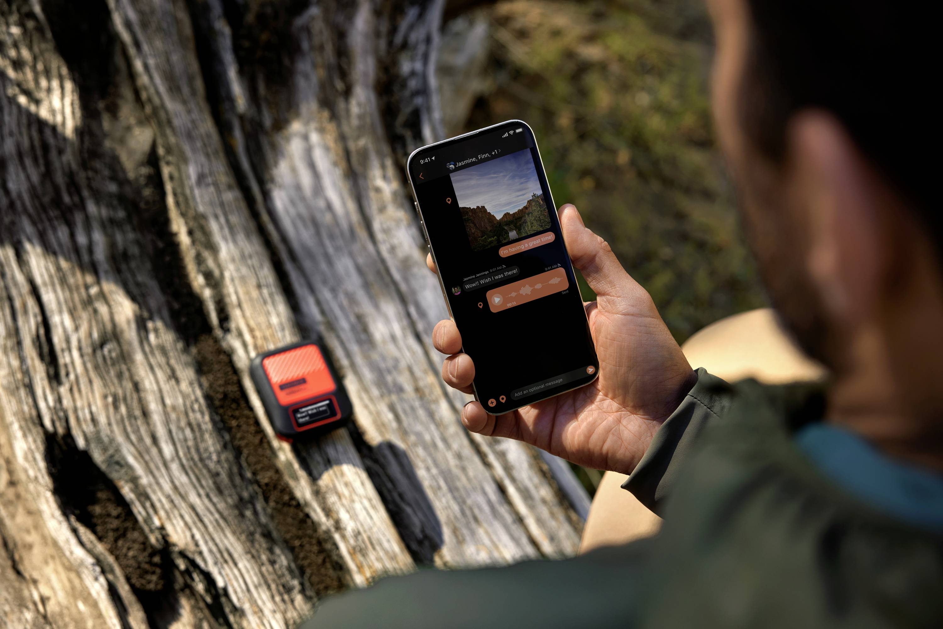A person is viewing an outdoor message on their smartphone, with a portable red and black device beside a tree trunk.
