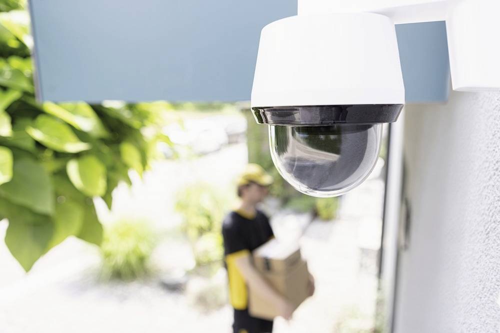 A security camera is in focus in the foreground, while in the background a person is delivering a package.