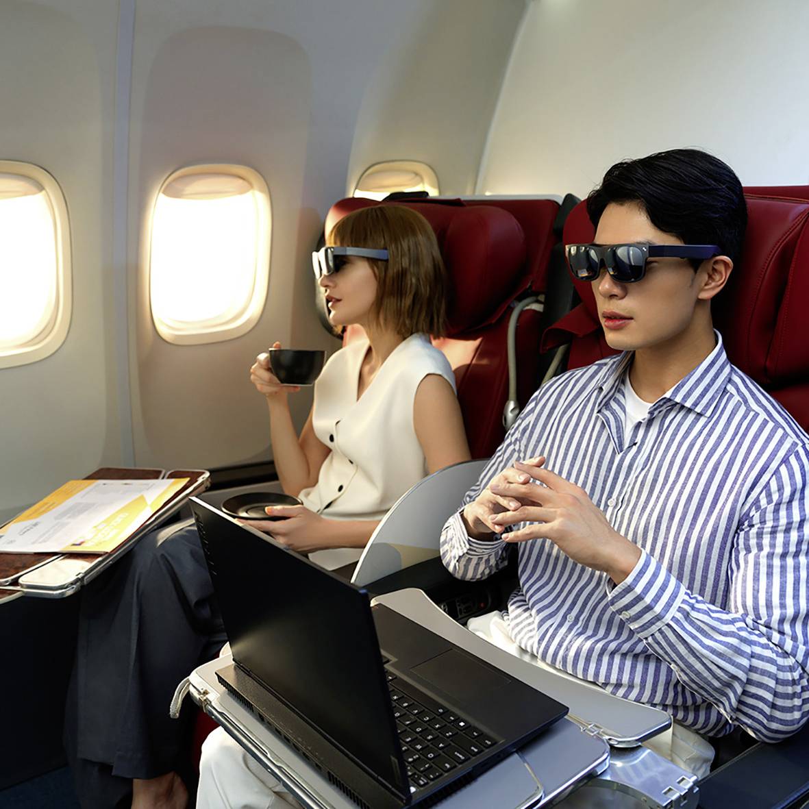 A man on the plane is wearing sunglasses and working on a laptop. A woman is sitting next to him, drinking from a paper cup.