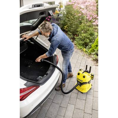 A man is cleaning the boot of a white car with a yellow hoover on a paved driveway beside a blooming garden.