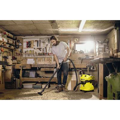 A man is using a vacuum cleaner in a workshop to remove wood shavings from the floor. Tools are visible in the background.