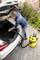 A man is cleaning the boot of a white car with a yellow vacuum cleaner on a paved driveway.