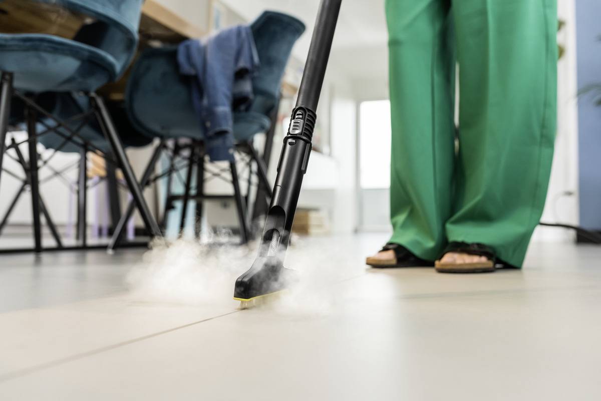 A person cleaning a tiled floor with a steam cleaner. Steam is visibly rising from the cleaner head. Furniture is in the background.