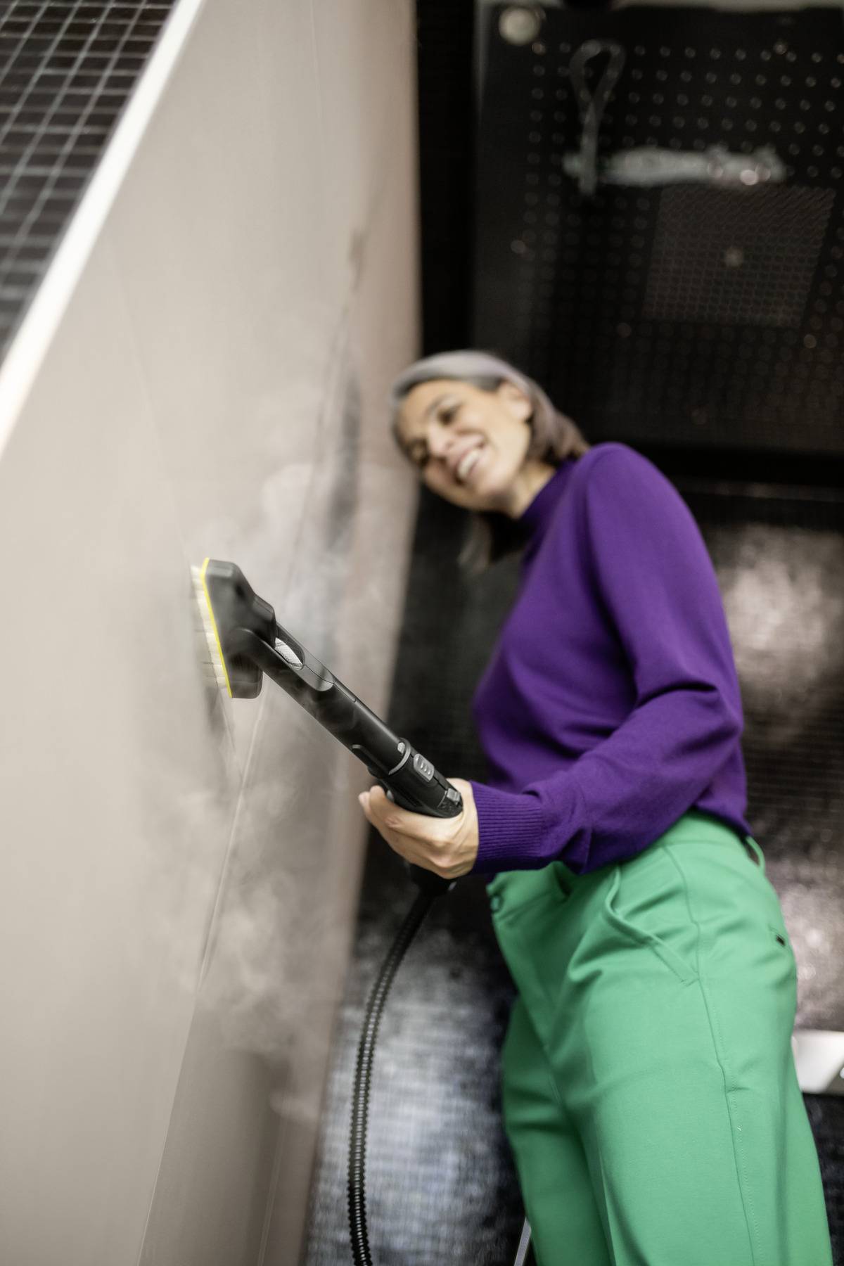 A person in a purple sweater and green pants uses a steam cleaner on a tiled wall, smiling slightly.
