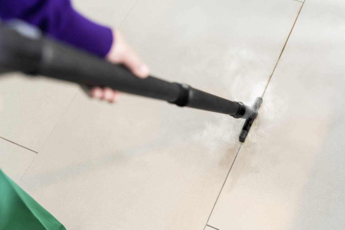 A person uses a steam cleaner on a tile floor, focusing on grout lines.