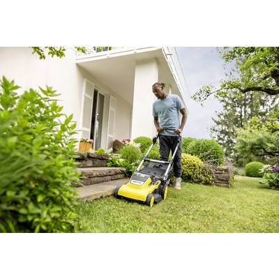 A man is mowing the lawn with a yellow lawnmower in front of a white house. The surroundings show flowering plants and green foliage.