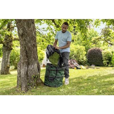 A person empties the grass collector of a lawnmower into a green garden bag beneath a tree in the garden.