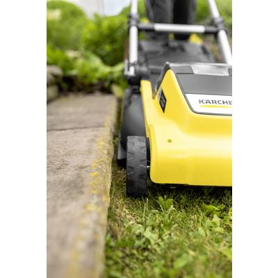 A lawnmower cuts grass along a garden path. Focus on the yellow machine and its wheels. Green plants in the background.