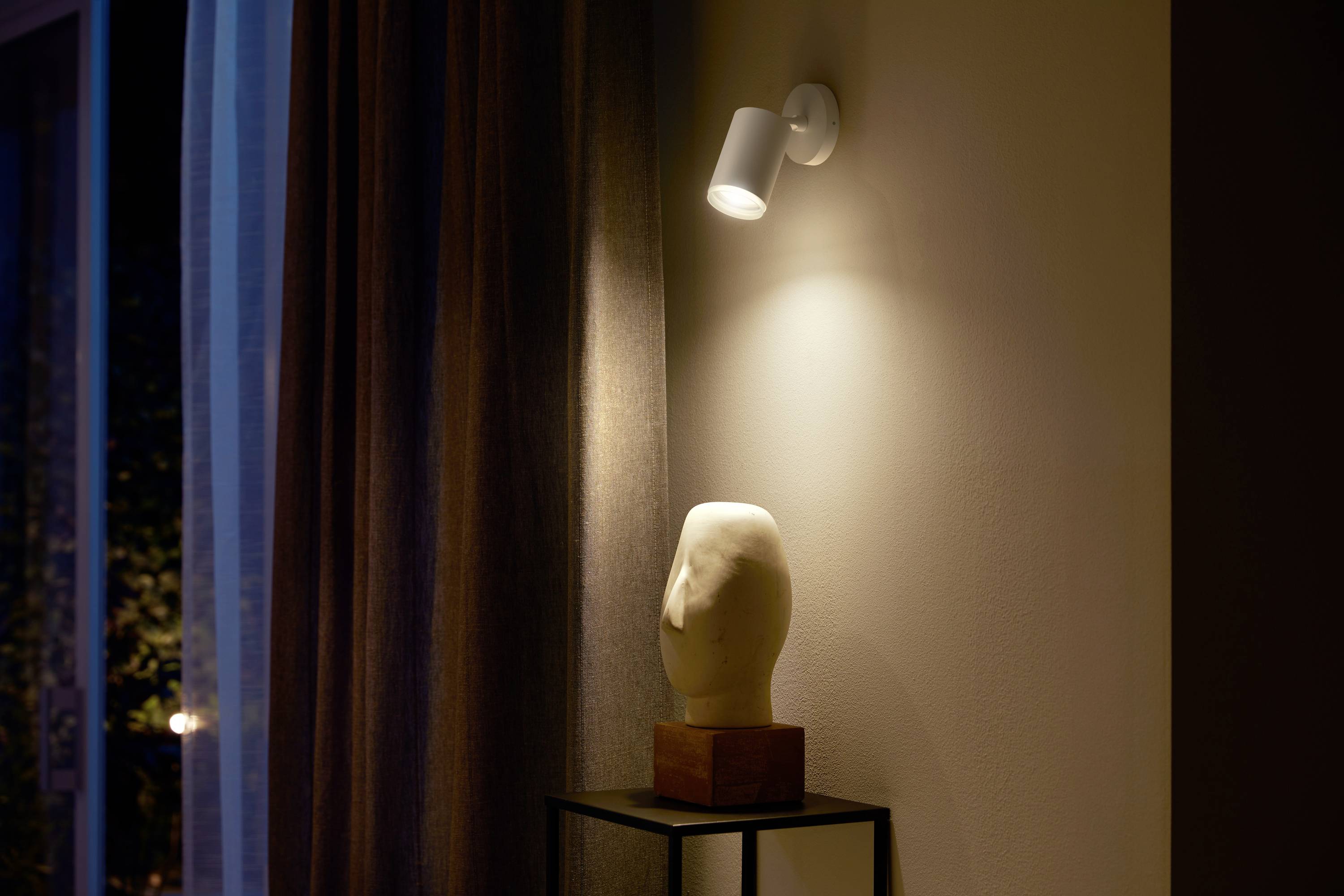 A lit room with a white mannequin head on a black plinth, illuminated by a spotlight. Curtains are visible in the background.