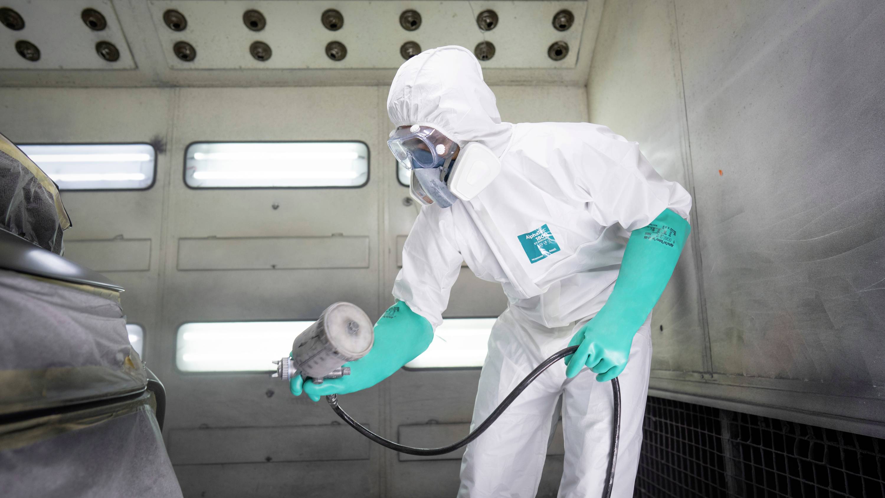 A person in a protective suit and respiratory mask is spray painting a car in a booth. They are wearing green gloves and holding a spray gun.