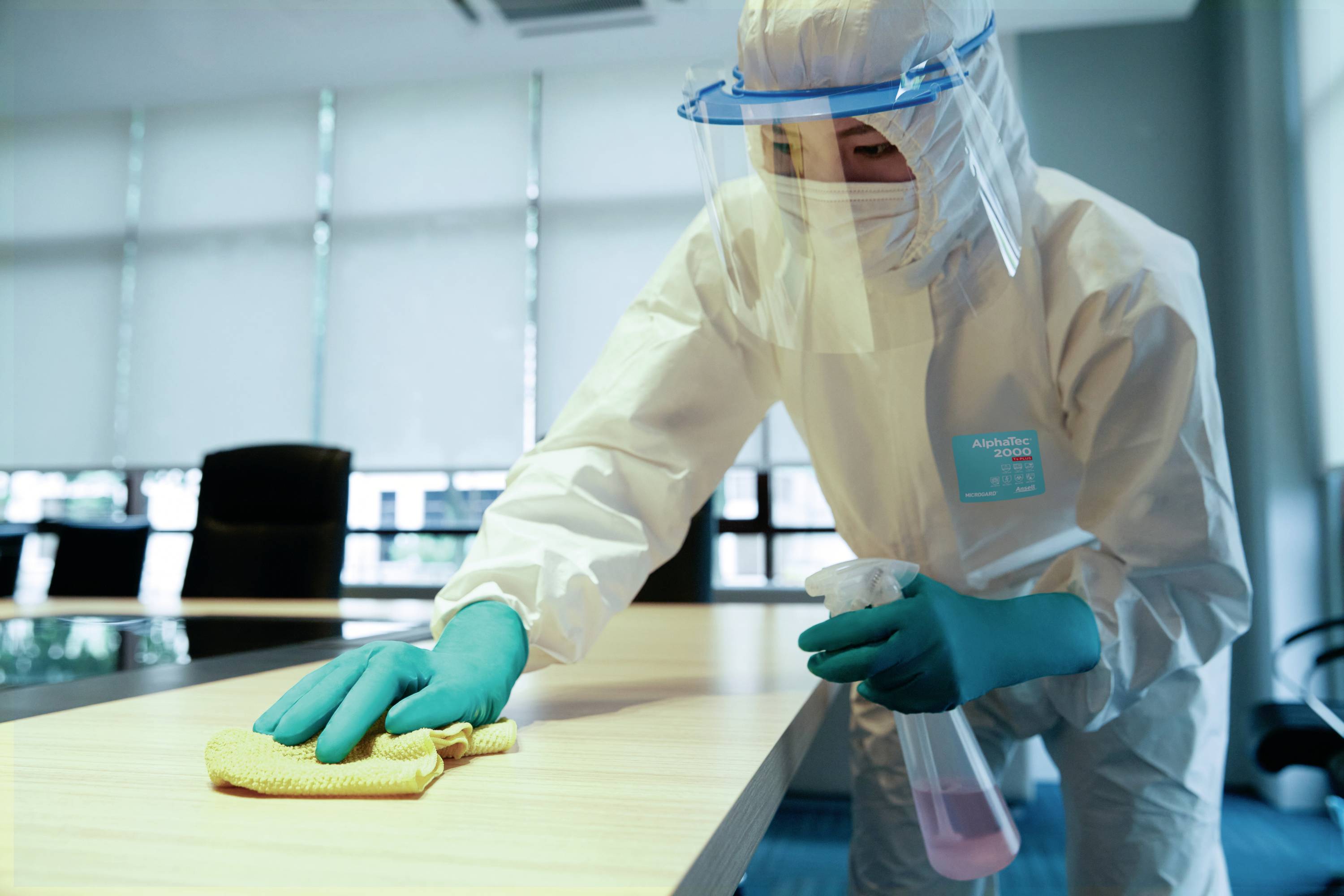 A person in protective clothing is disinfecting a table in an office.
