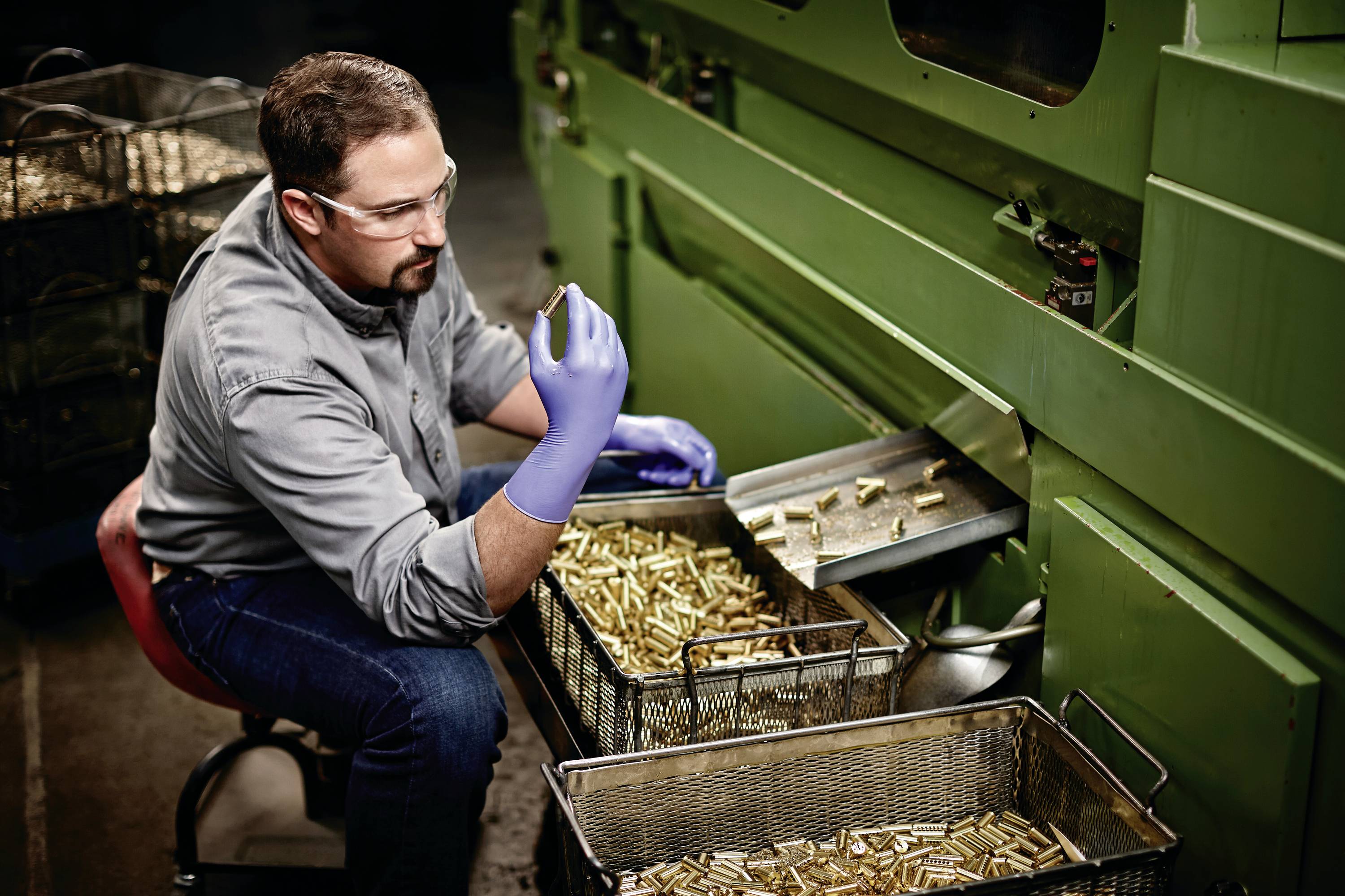 A man is inspecting cartridge casings in a factory. He is wearing safety glasses and gloves. Baskets full of casings are standing next to him.
