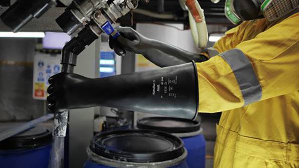 A person wearing yellow protective clothing and black gloves is operating a valve near large blue barrels in an industrial setting.