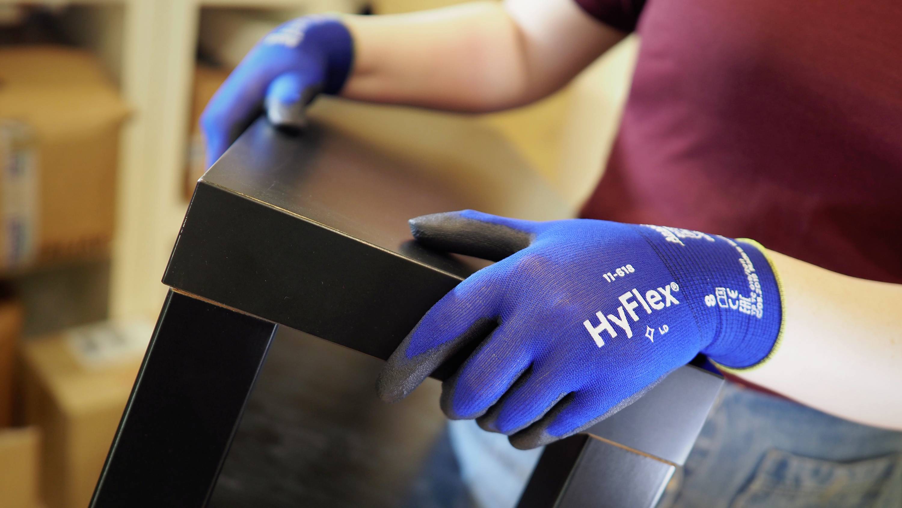 A person wearing blue gloves is holding and moving a table leg. Cardboard boxes and a shelf are visible in the background.