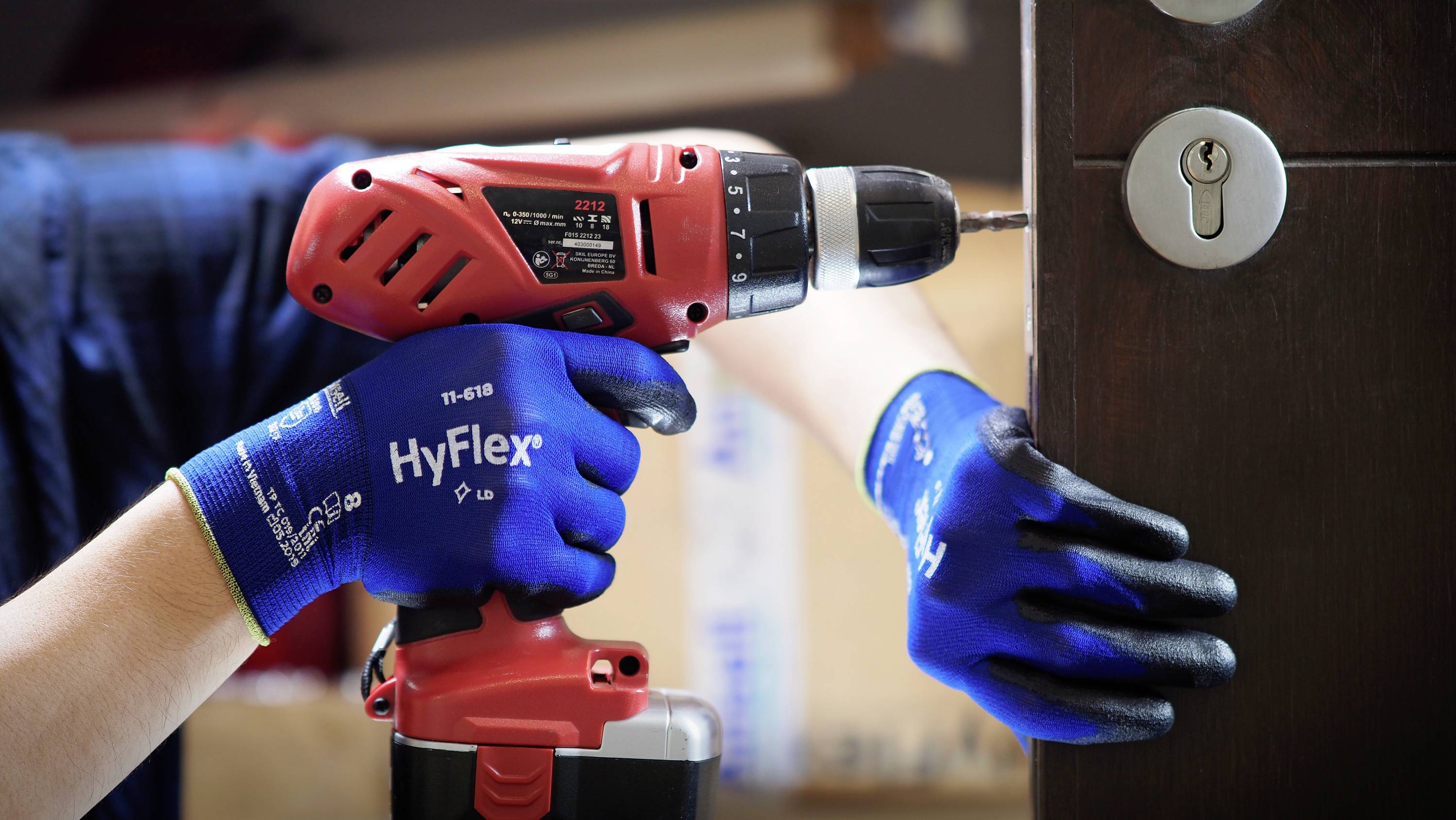 A person is drilling a hole in a wooden door using a red cordless drill. They are wearing blue work gloves. A keyhole is visible.
