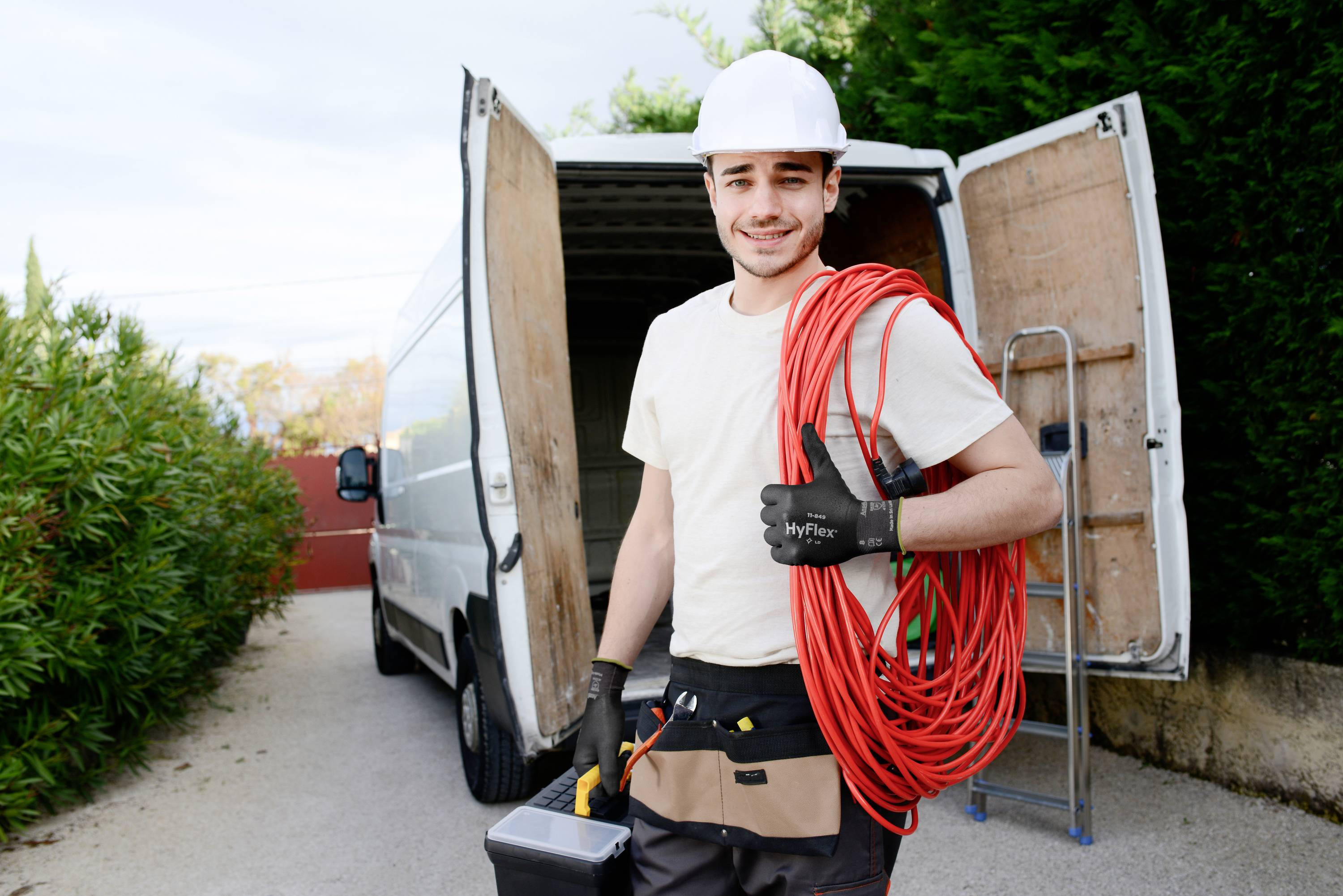 A tradesman in work attire stands in front of an open van, holding a long red cable and carrying a tool bag.