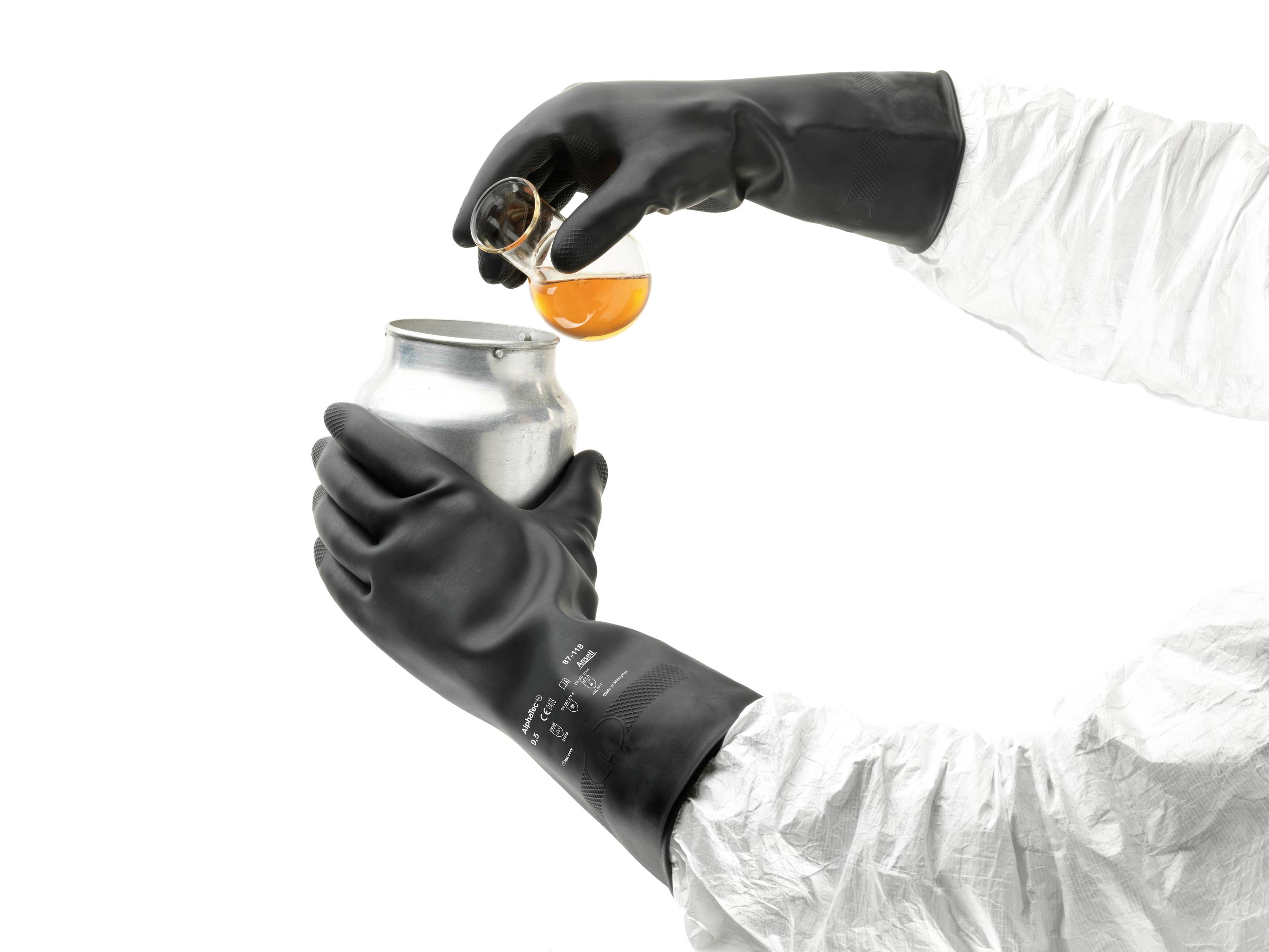 A person wearing protective equipment is pouring orange liquid from a beaker into a metal container. Hands are wearing black gloves.