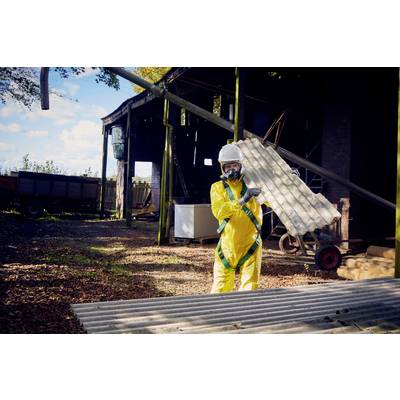 A person wearing a yellow protective suit and respirator transports corrugated sheets on an industrial site during daylight.