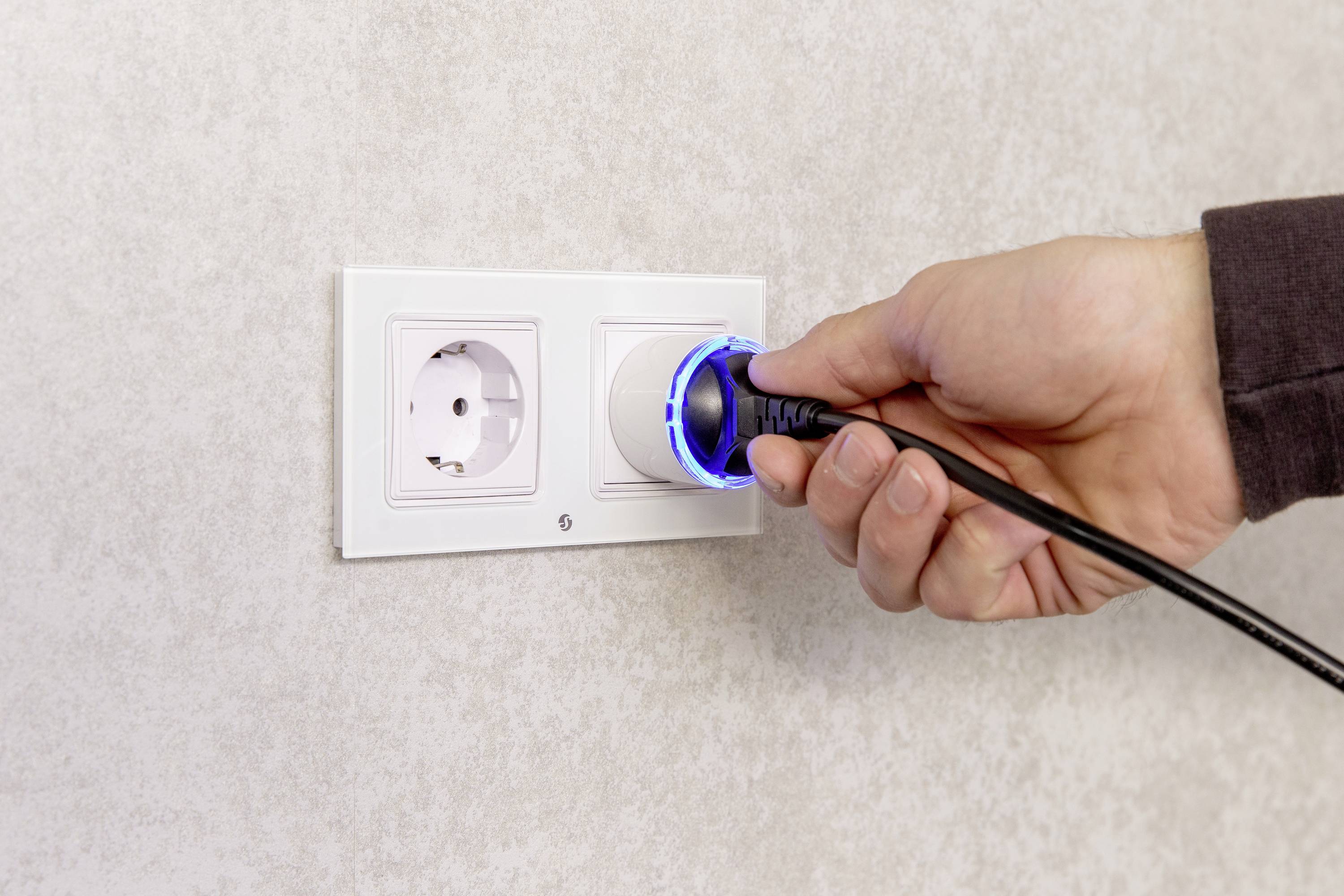 A hand is plugging a plug into a white socket on a wall.