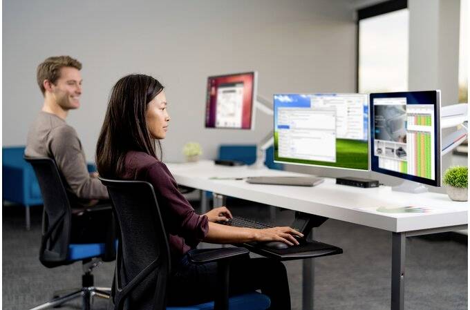 Two people are working intently on computers in a modern office. One is looking at multiple screens, the other is smiling.