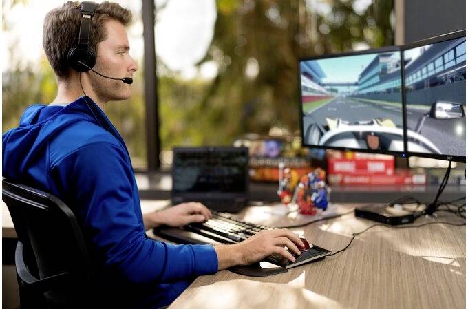 A man sits at a desk, wearing a headset and playing a racing video game on two monitors. Trees are visible in the background.