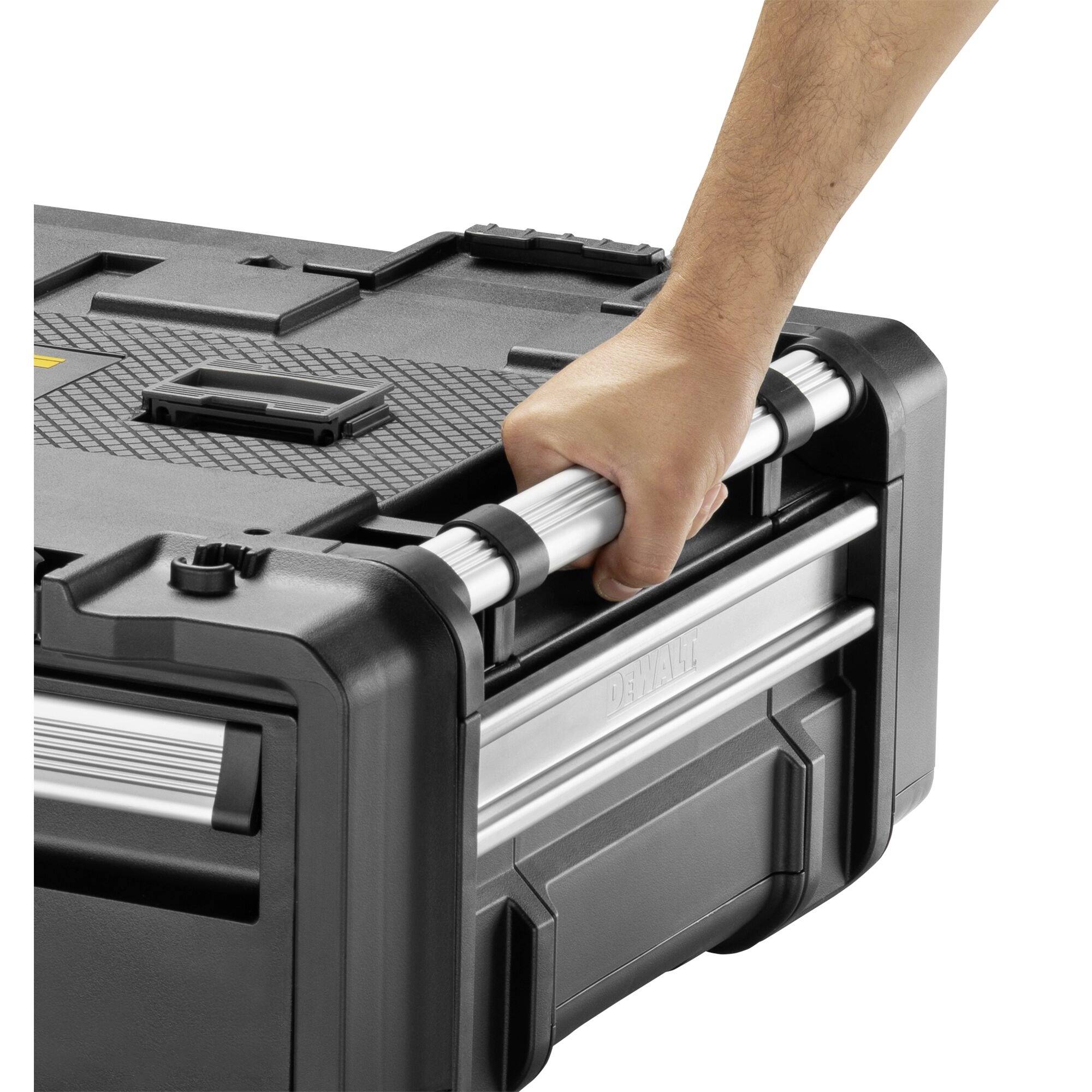 A hand lifts a black tool box with a silver handle. The box has a robust construction and textured surface.