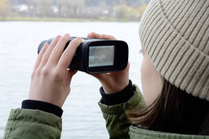 A person is viewing the landscape by the water through a pair of binoculars; winter clothing suggests cold weather.