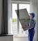 A worker in a blue overall is installing a window pane in a modern room with white curtains.