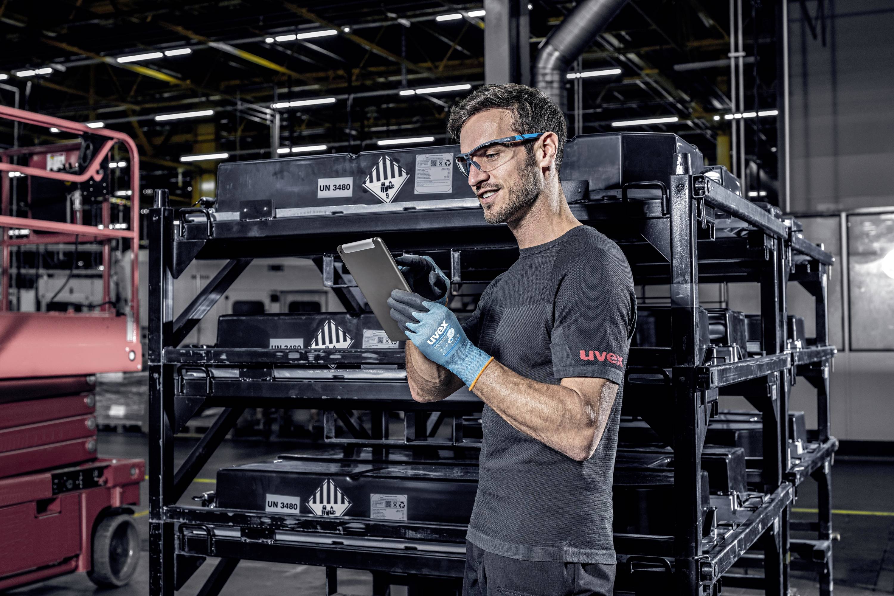 A warehouse worker wearing safety glasses and gloves is using a tablet and standing in front of shelves stacked with boxes.