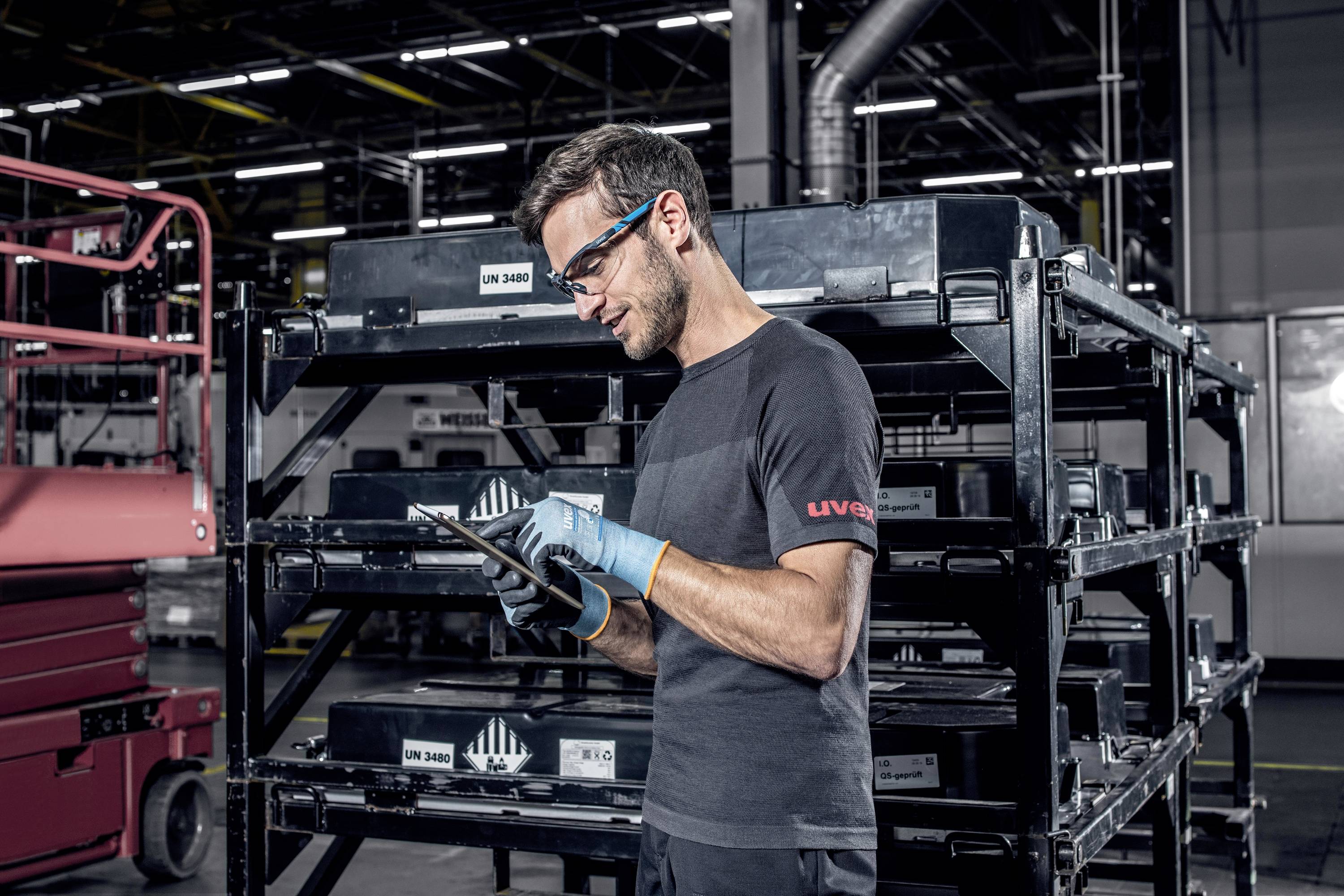 A man in workwear and safety glasses is standing in a factory hall, checking information on a tablet in front of black shelves with crates.