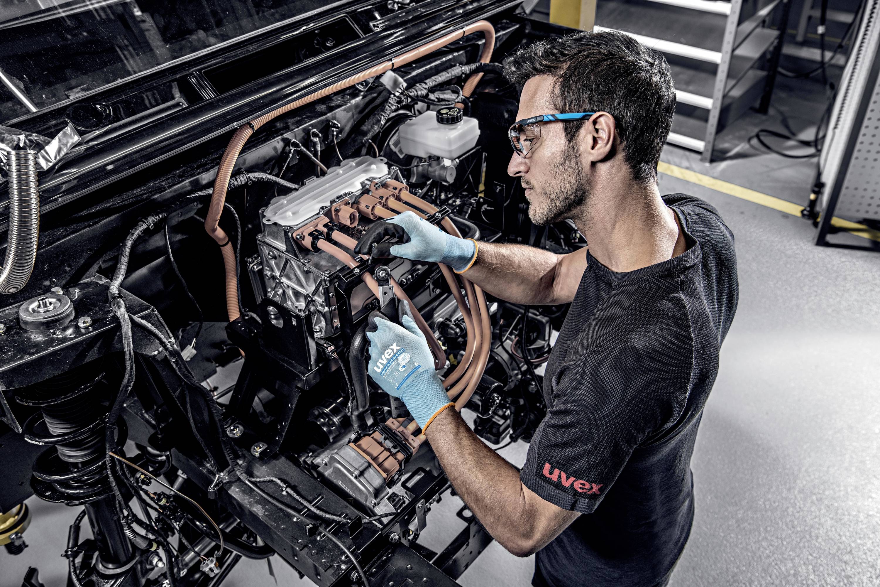 A man wearing safety glasses and gloves is working on a car electric motor in a workshop.