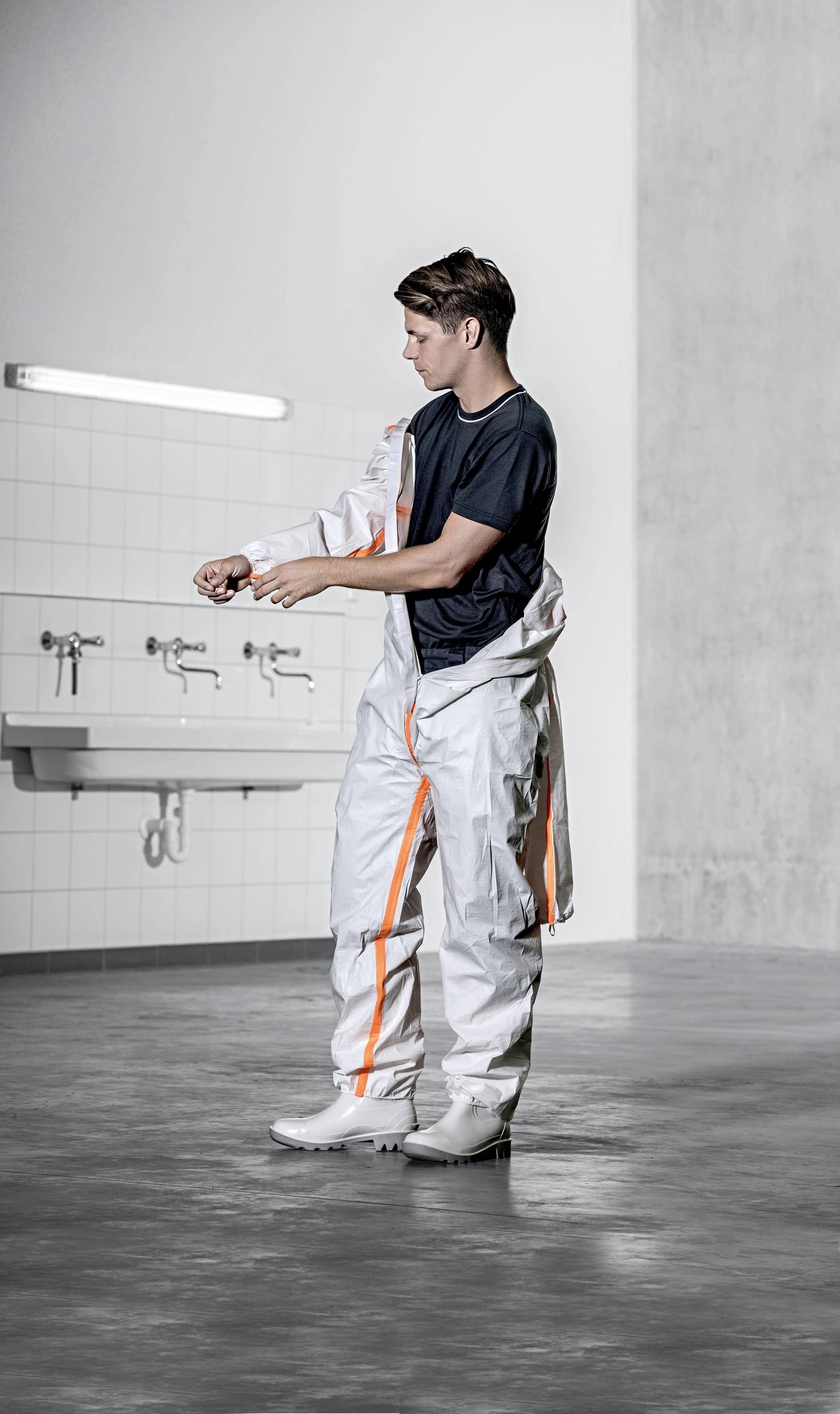 A person is putting on a white protective suit with orange stripes in an industrial space. Sinks can be seen in the background.