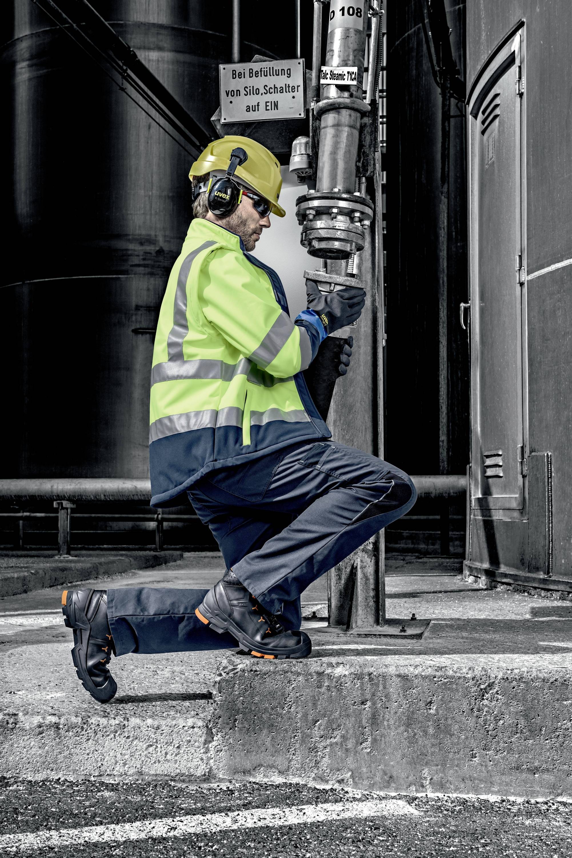A worker in safety clothing is kneeling to operate a valve in an industrial facility. Pipes and signs are visible in the background.