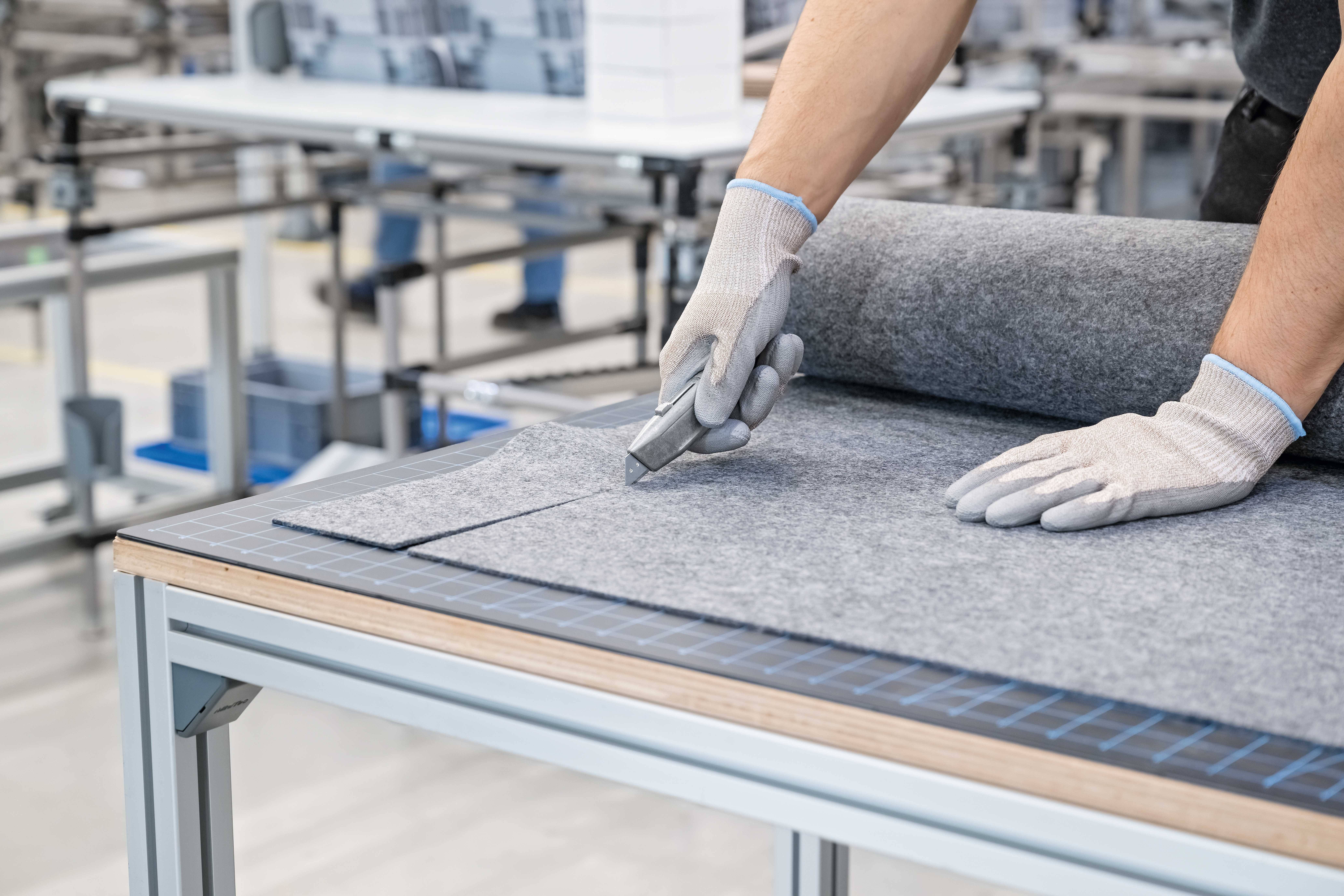 A person is cutting grey felt fabric on a table using a craft knife. The hands are wearing white work gloves.
