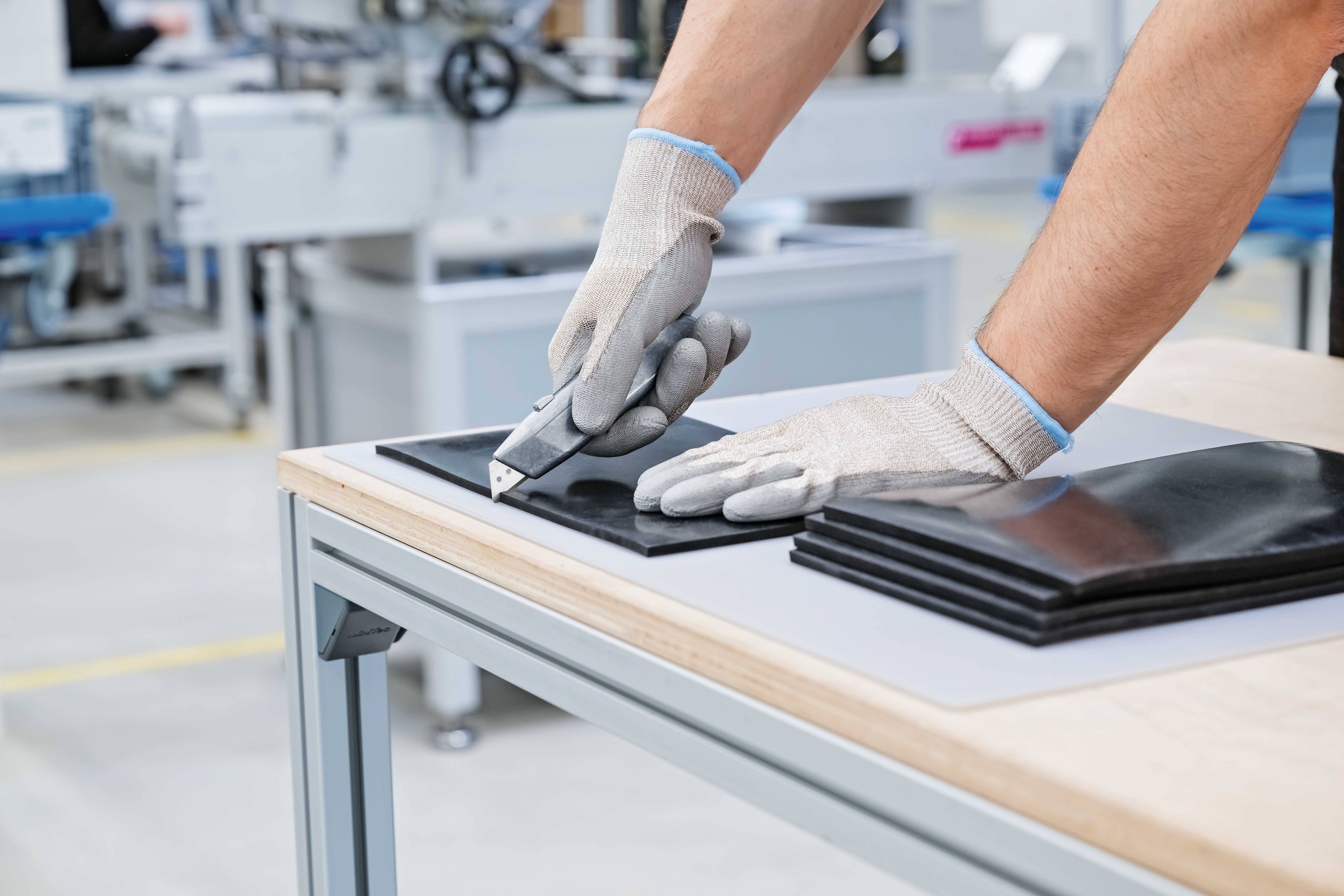 A person is cutting black rubber sheets with a craft knife on a wooden table. They are wearing grey gloves.