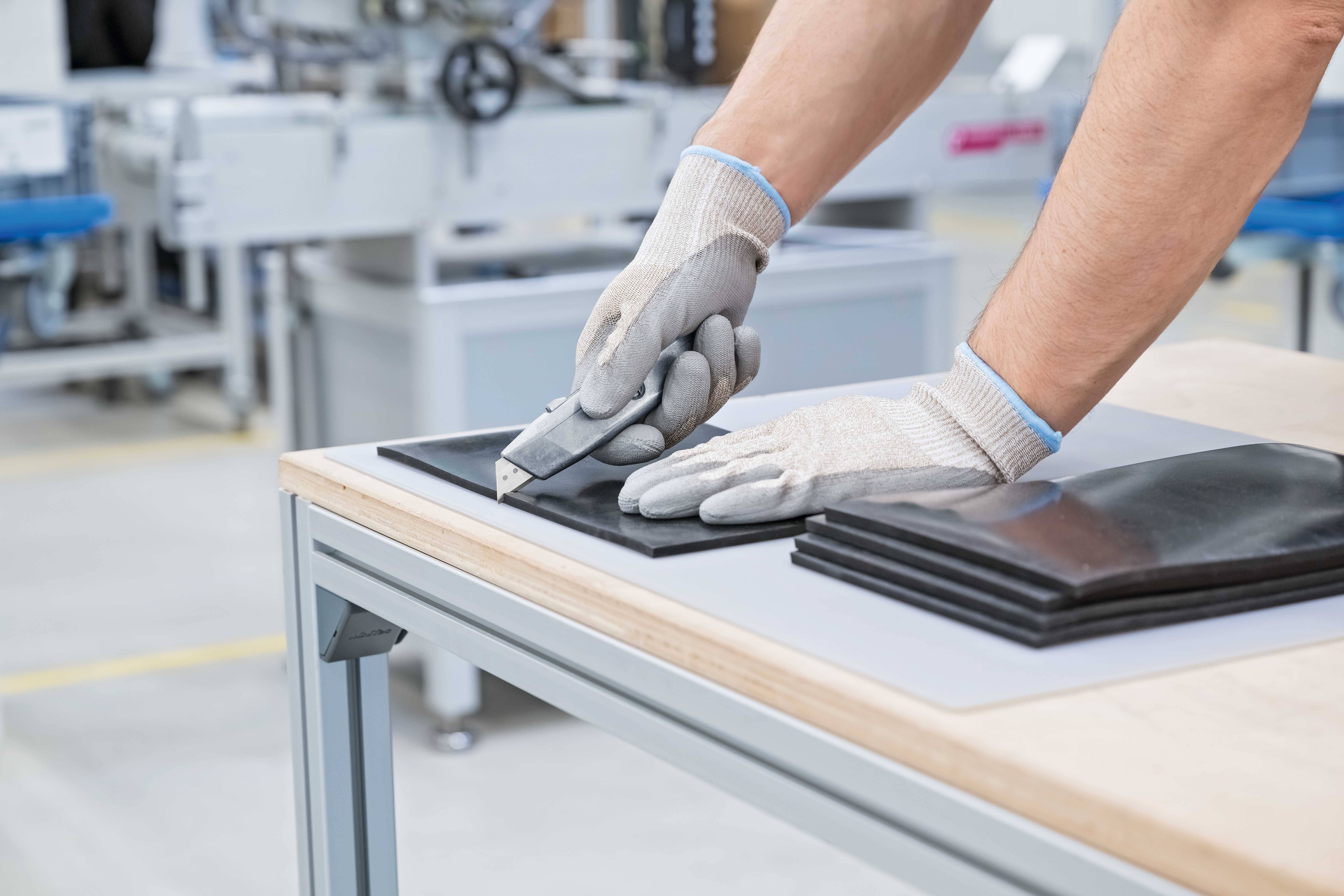 A worker wearing gloves is cutting rubber material with a knife on a table in a factory environment.
