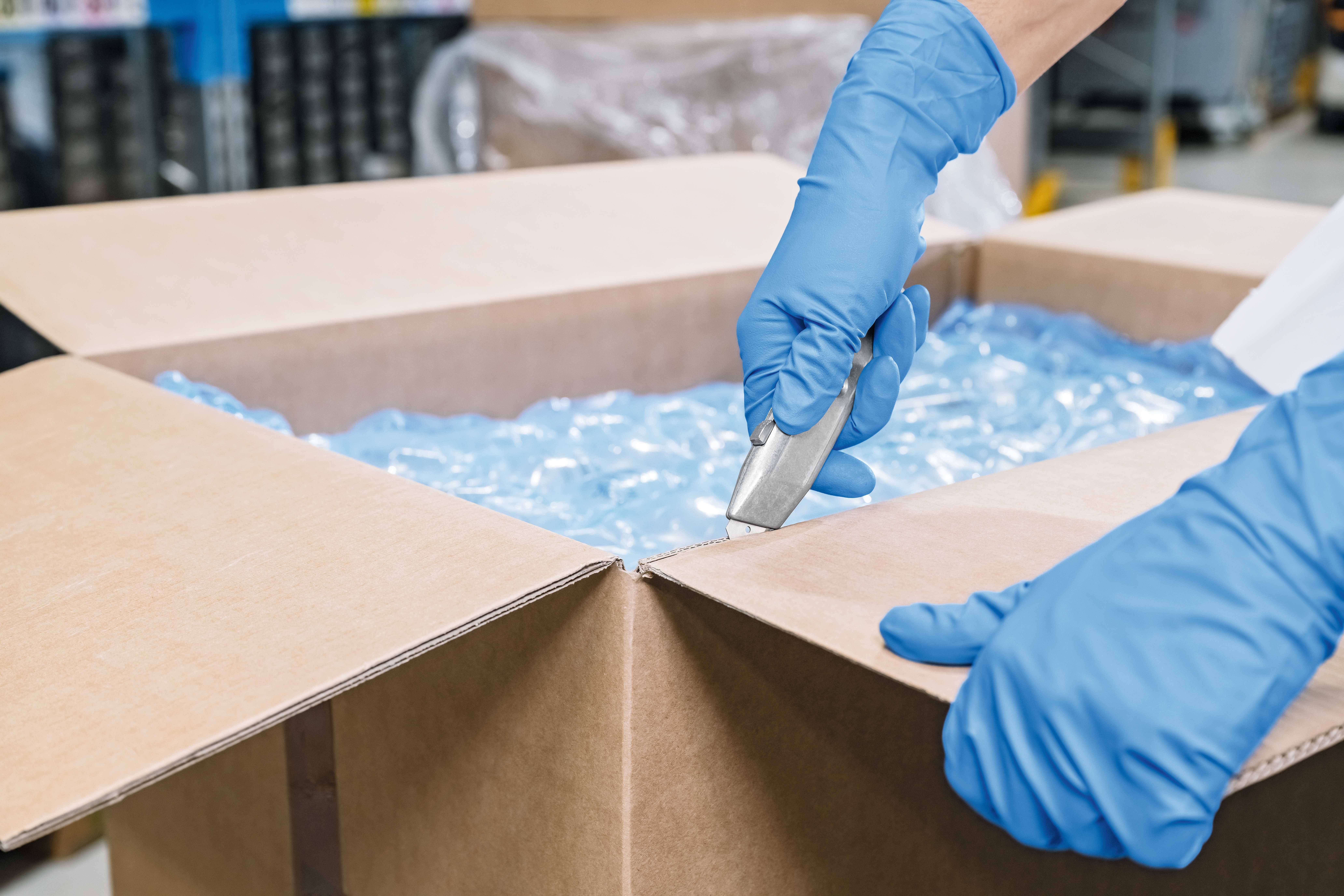 A person wearing blue gloves carefully cuts a cardboard box with a utility knife. Packaging material is visible.