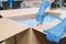 A person wearing blue gloves carefully cuts a cardboard box with a utility knife. Packaging material is visible.