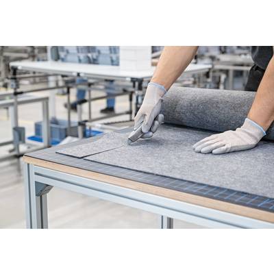 A person wearing gloves is cutting grey fabric on a table in a factory environment. The focus is on precise manual work.